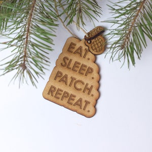May include: A wooden Christmas ornament shaped like a tree with the text "Eat. Sleep. Patch. Repeat."  The ornament is decorated with a small wooden pine cone charm.