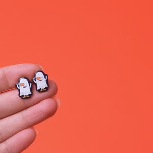 May include: A pair of white ghost earrings with black outlines and orange eyes. The earrings are on a hand against a bright orange background.