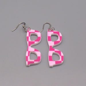 Pode incluir: Um par de brincos pendentes com um design plano e recortado em forma de óculos. Os brincos apresentam um padrão xadrez rosa vibrante e branco em toda a superfície e estão presos a ganchos de metal de tom prateado.