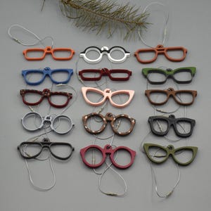 以下が含まれることがあります： オレンジ、白、茶色、青、ピンク、緑など、さまざまな形と色のカラフルなメガネオーナメントのコレクション。各オーナメントには、吊り下げ用の銀色の紐が付いています。オーナメントは灰色の表面に配置されています。