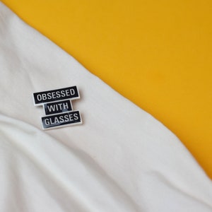 May include: A rectangular enamel pin with the text "OBSESSED WITH GLASSES" in black letters on a white background. The pin is on a white fabric with a yellow background.