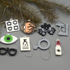 Puede incluir: Una colección de adornos con temática de ojos, que incluyen una tabla optométrica, gafas y un ojo con iris verde. Otros adornos incluyen un ojo, una máquina de examen ocular y un foróptero. Los adornos son blancos, negros, grises y beige.