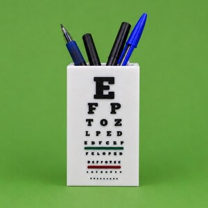 May include: A white rectangular pen holder featuring a black Snellen eye chart design on the front. The holder contains four pens in blue and black ink colors. The eye chart includes rows of letters in decreasing size with horizontal lines.