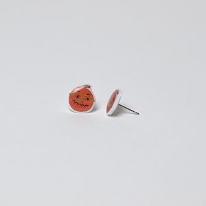 May include: A pair of orange pumpkin stud earrings with a black smiling face and gold glasses. The earrings are on a white background.