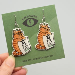 Pode incluir: Um par de brincos pendentes de olho de gato laranja com um gancho prateado. O gato está segurando um cartão de visão que diz "E F P T O C" e "L P E D P E F O Z". Os brincos estão em um cartão verde com o texto "Eye Power" e um raio.