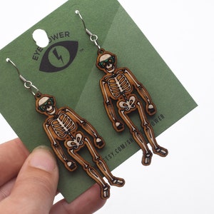 Könnte beinhalten: Ein Paar Skelett-Ohrringe aus Holz mit grünen Sonnenbrillen. Die Ohrringe hängen an silbernen Haken und befinden sich auf einem grünen Hintergrund mit dem Text "Eye Power" darauf.