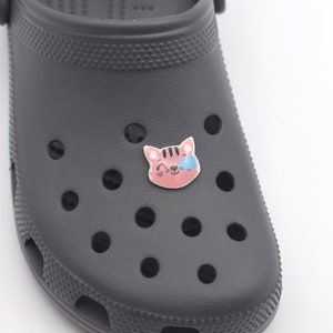 May include: A black clog with a pink and blue cat charm. The cat charm has a winking eye and a pink nose.