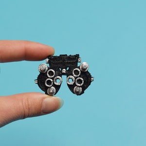 Könnte beinhalten: Ein Miniatur-Phoropter in Schwarz, ein Gerät, das bei Augenuntersuchungen verwendet wird, mit silberfarbenen Drehknöpfen und Linsenhaltern. Das Gerät wird vor einem hellblauen Hintergrund gehalten und zeigt sein kompliziertes Design und seine geringe Größe.