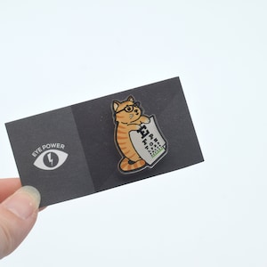 May include: A black card with the text "Eye Power" and a white eye with a lightning bolt inside. The card is holding a clear plastic pin of a cartoon orange cat wearing glasses and holding an eye chart.