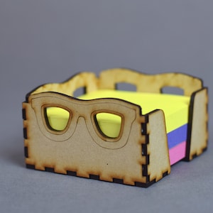 Könnte beinhalten: Holz-Notizblockhalter mit einem ausgeschnittenen Brillenpaar an der Vorderseite. Der Halter ist mit gelben, blauen und rosa Haftnotizen gefüllt.