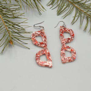 Pode incluir: Um par de brincos vermelhos de glitter em forma de óculos. Os brincos estão pendurados em ganchos prateados.