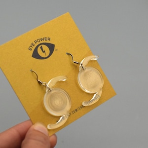 May include: A pair of clear acrylic earrings with a spiral design, displayed on a mustard yellow card. The card features the text "EYE POWER" and a graphic of an eye with a lightning bolt. The earrings have silver-colored hooks.