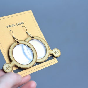 Pode incluir: Um par de brincos de madeira em forma de lupas com um gancho dourado. Os brincos têm uma lente de plástico transparente e uma alça de madeira com o texto "Trial Lens" e os números " +2" e " +3" impressos neles.
