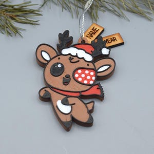 May include: A wooden Christmas ornament in the shape of a reindeer. The reindeer has a red scarf, a Santa hat, and a white and red polka-dotted cheek. The ornament has two small wooden tags that read "NAME" and "YEAR".