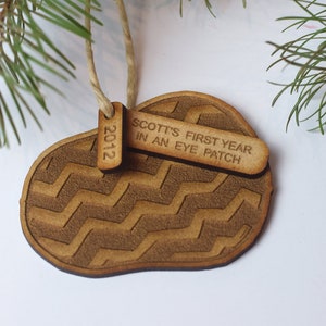 May include: A wooden Christmas bauble shaped like a footprint with a zig-zag pattern. It has two small wooden tags attached to it, one that says "2012" and the other that says "SCOTT'S FIRST YEAR IN AN EYE PATCH".