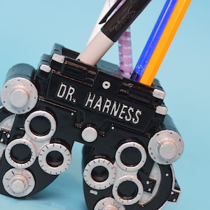 May include: Black eye chart shaped pen holder with silver accents. The holder has the text "DR. HARNESS" on the front. Three pens are in the holder.