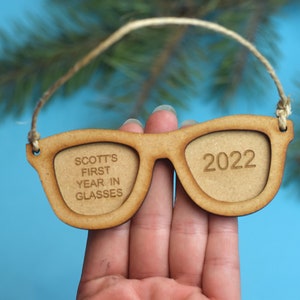 以下が含まれることがあります： 片方のレンズに「SCOTT'S FIRST YEAR IN GLASSES」、もう片方のレンズに「2022」と刻まれた、サングラスの形をした木製オーナメントです。