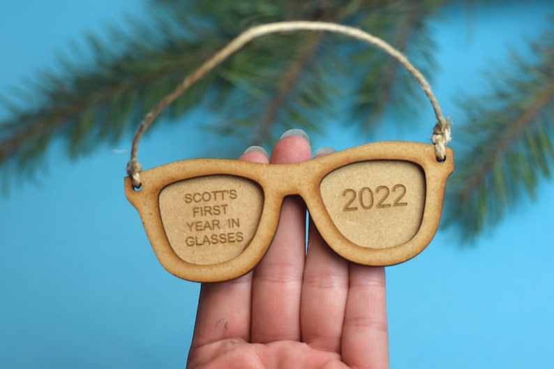 以下が含まれることがあります： 片方のレンズに「SCOTT'S FIRST YEAR IN GLASSES」、もう片方のレンズに「2022」と刻まれた、サングラスの形をした木製オーナメントです。