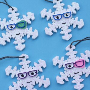 May include: Four white glitter snowflake ornaments with black string hangers. Each snowflake has a different colored pair of glasses and a smiley face.