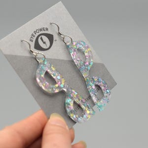 Pode incluir: Um par de brincos de acrílico transparente com glitter em forma de óculos, com glitter iridescente suspenso no interior. Os brincos penduram-se de ganchos prateados e são exibidos num cartão cinzento com o texto "EYE POWER".