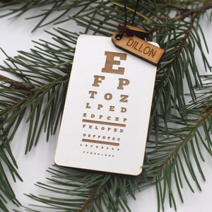 May include: A white wooden eye chart ornament with the letters E, FP, TOZ, LPED, EDFCZP, FELOPZD, DEFOTEC, LEFODP, and FYRNAIDEC printed on it. The ornament has a small wooden tag attached to it that says "DILLON".