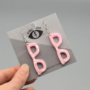 Pode incluir: Brincos em forma de óculos rosa com um padrão de redemoinho branco. Os brincos estão pendurados em ganchos prateados. Os brincos estão em um cartão cinza com o texto "EYE*NER" e um logotipo de raio.