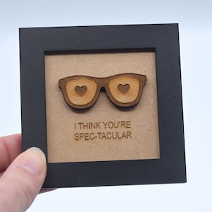 May include: A wooden framed sign with a laser cut design of glasses with hearts in the lenses. The sign reads "I think you're spec-tacular".