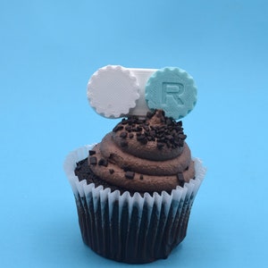 May include: A chocolate cupcake with chocolate frosting and sprinkles topped with a white and blue 3D printed cupcake topper with the letter "R" on it.