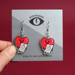 May include: A pair of red heart-shaped earrings with a cartoon heart holding an eye chart. The earrings are silver and have a hook closure. The earrings are on a gray card with the text "Eye Power" and a lightning bolt logo.