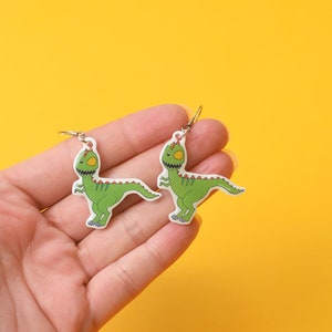 Pode incluir: Um par de brincos de dinossauro verde com ganchos prateados. Os dinossauros estão olhando em direções opostas.