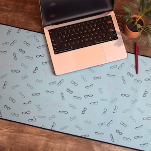 Pode incluir: Um tapete de mesa azul claro com um padrão repetitivo de óculos pretos. O tapete está sobre uma superfície de madeira com um computador portátil e uma planta em vaso.