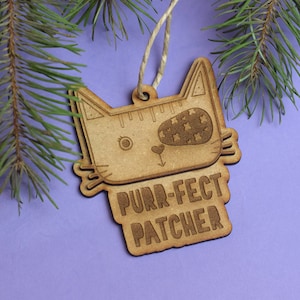 May include: A wooden cat-shaped ornament with the text "Purr-fect Patcher" hanging from a string against a purple background.