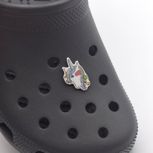 Pode incluir: Um tamanco preto com um amuleto de unicórnio branco preso na frente. O amuleto tem uma crina e uma cauda arco-íris.