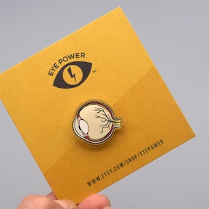 May include: A gold-colored enamel pin featuring a detailed anatomical illustration of a human eye. The pin is on a yellow card with the text "EYE POWER" and a lightning bolt graphic. The card also includes the website address "www.etsy.com/shop/EYEPOWER".