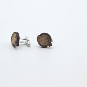 May include: A pair of wooden cufflinks with a detailed anatomical eye design. The cufflinks are brown with a light brown eye and are attached to silver metal cufflink backs.