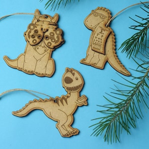 May include: Three wooden ornaments shaped like dinosaurs. One dinosaur is wearing glasses and holding an eye chart, another is wearing glasses and looking through a magnifying glass, and the third is smiling with its mouth open. The ornaments are on a blue background.