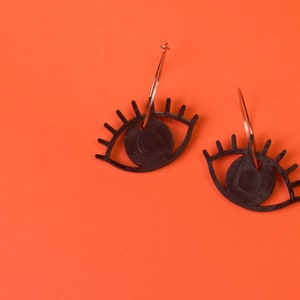 Pode incluir: Um par de brincos pretos em forma de olho com argolas douradas. Os brincos estão sobre um fundo laranja brilhante.