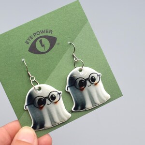 May include: Ghost-shaped earrings with black-rimmed glasses and smiling faces. The earrings are displayed on a green card with the text "EYE POWER" and an eye logo. The earrings hang from silver-coloured fish hook ear wires.
