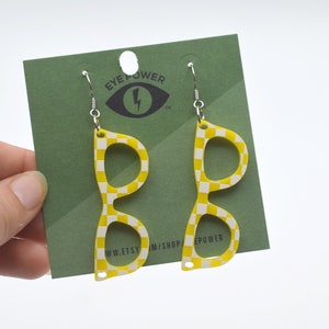 Pode incluir: Brincos em forma de óculos quadriculados amarelos e brancos com ganchos prateados. Os brincos estão em um cartão verde com o texto "Eye Power" e um raio.