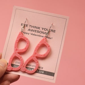 Pode incluir: Brincos de óculos de coração rosa com ganchos prateados. Os brincos estão em um cartão rosa com o texto "EYE THINK YOU'RE AWESOME Happy Valentine-day!" e o texto "ETSY EYE KIDS B.COM SHOP/EY! WER".