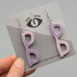 Pode incluir: Um par de brincos de óculos com gradiente de cor roxo e rosa com ganchos prateados. Os brincos são exibidos em um cartão com o texto "EYE POWER" e um logotipo de raio.