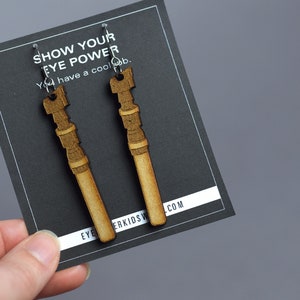 Pode incluir: Um par de brincos de madeira em forma de sabres de luz. Os brincos são marrons e têm um gancho prateado. O cartão atrás dos brincos diz "Show Your Eye Power You have a cool job." e "eyekidsw.com".