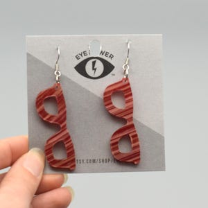 Pode incluir: Um par de brincos em forma de óculos listrados em vermelho e branco com ganchos prateados. Os brincos são exibidos em um cartão cinza com o texto "EYE NER" e um logotipo de raio. O URL da loja Etsy também é visível.