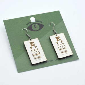 Pode incluir: Um par de brincos de madeira em forma de tabela de optometrista com ganchos prateados. Os brincos são retangulares e apresentam uma tabela de optometrista clássica com as letras E, F, P, T, O, Z, L, P, E, D, R, e algumas linhas de letras menores. Os brincos estão sobre um cartão verde com as palavras "Eye Power" e um olho estilizado com um raio.