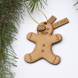 May include: Wooden gingerbread man ornament with a brown and white icing design. The ornament has a string for hanging and a tag that says "YEAR" and "YOUR NAME".