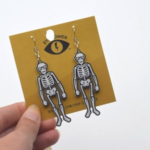 Könnte beinhalten: Ein Paar Skelett-Ohrringe mit Brille. Die Ohrringe sind silberfarben und haben einen schwarzen Umriss. Die Ohrringe befinden sich auf einer gelben Karte mit dem Text "Eye Power" und einem Blitz.