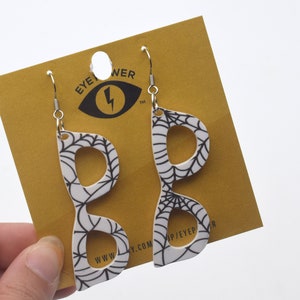 Pode incluir: Um par de brincos brancos pendurados em forma de óculos com um design de teia de aranha preta. Os brincos estão em um cartão amarelo com o texto "EYE POWER" e um logotipo de raio.