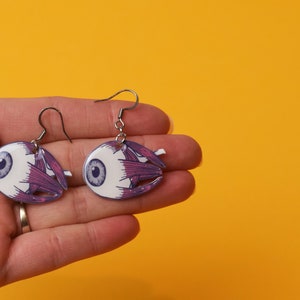 Pode incluir: Um par de brincos de prata com um design detalhado de olho anatômico. Os olhos são brancos com tecido muscular roxo e uma íris azul.