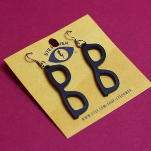 Pode incluir: Brincos pretos em forma de óculos em um cartão amarelo com o texto "EYE POWER" e "WWW.ETSY.COM/SHOP/EYEPOWER".