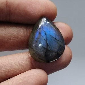 May include: A teardrop-shaped labradorite gemstone with a polished surface. The stone displays a mix of dark gray and blue hues, with flashes of iridescent blue light. The gemstone is held in a hand.